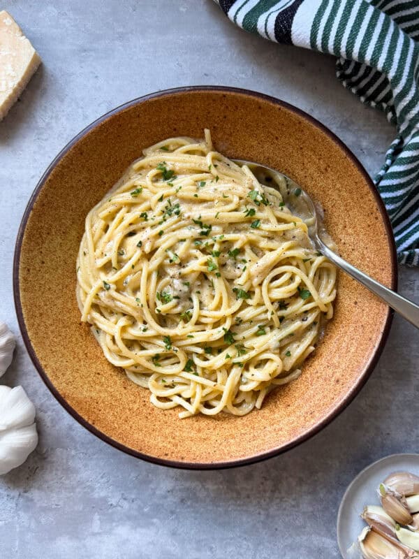 A bowl of creamy garlic pasta garnished with chopped herbs sits on a gray surface, with a fork in the dish. Nearby are a wedge of cheese, garlic cloves, and a striped kitchen towel—perfect inspiration for your next Garlic Pasta Recipe.