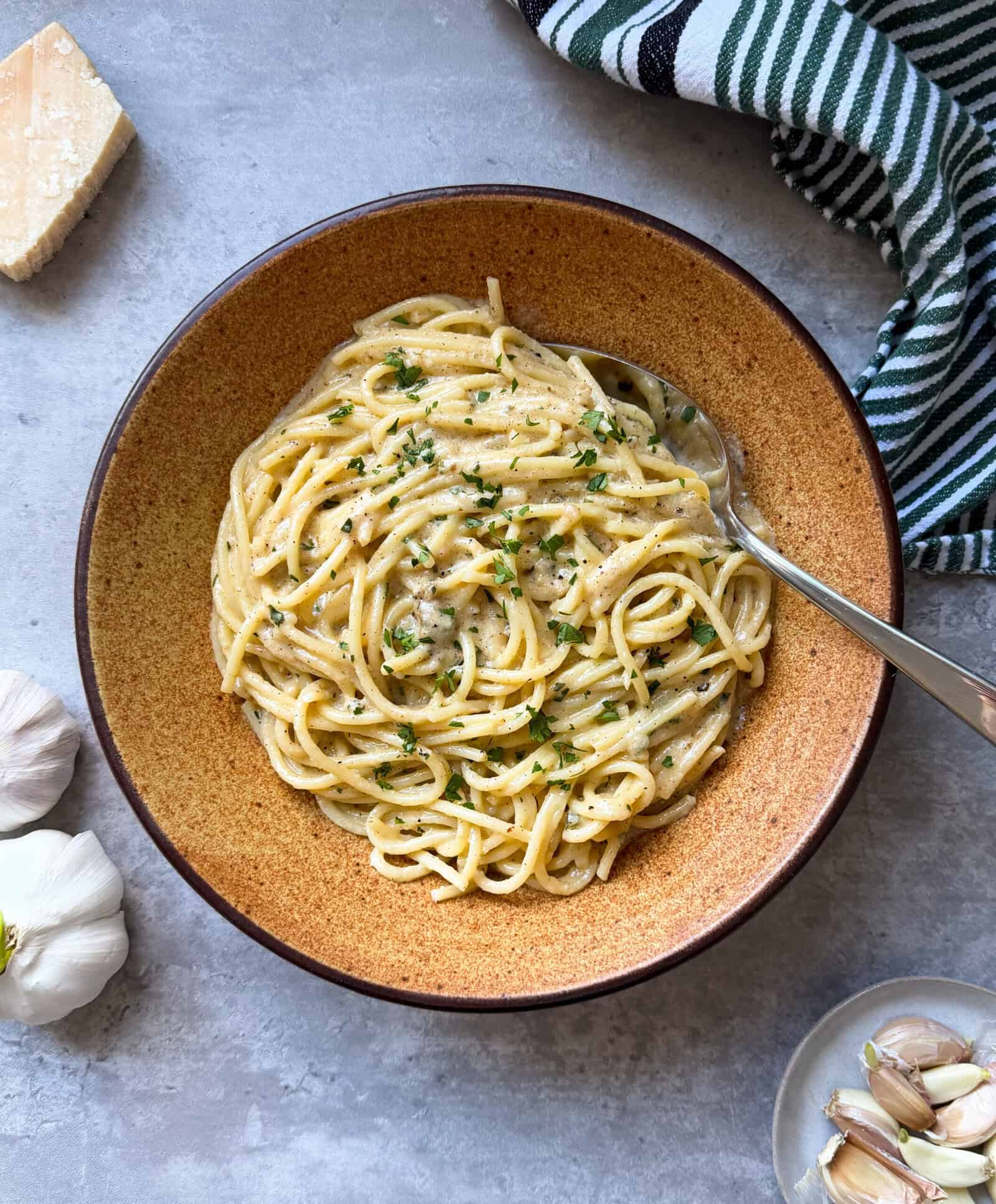 A bowl of creamy garlic pasta garnished with chopped herbs sits on a gray surface, with a fork in the dish. Nearby are a wedge of cheese, garlic cloves, and a striped kitchen towel—perfect inspiration for your next Garlic Pasta Recipe.