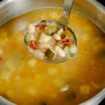 A metal ladle lifts chunky vegetable soup with white beans, zucchini, and pieces of tomato from a pot on a stove.