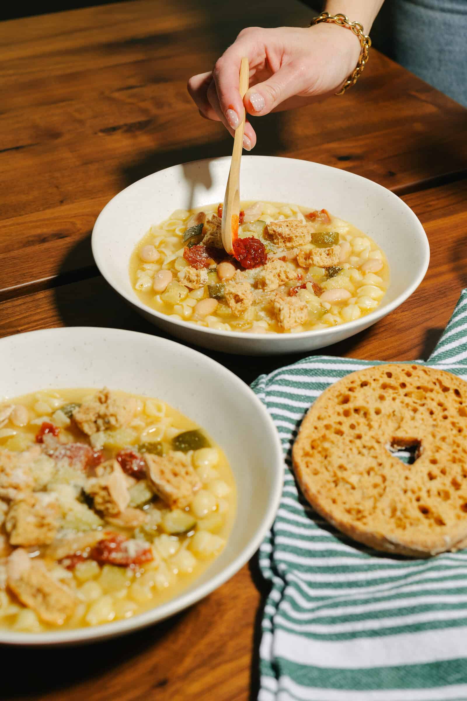 Two bowls of white bean soup with vegetables and shredded chicken sit on a wooden table; a hand lifts a spoonful from one bowl. A striped napkin and a toasted round bread are also on the table.