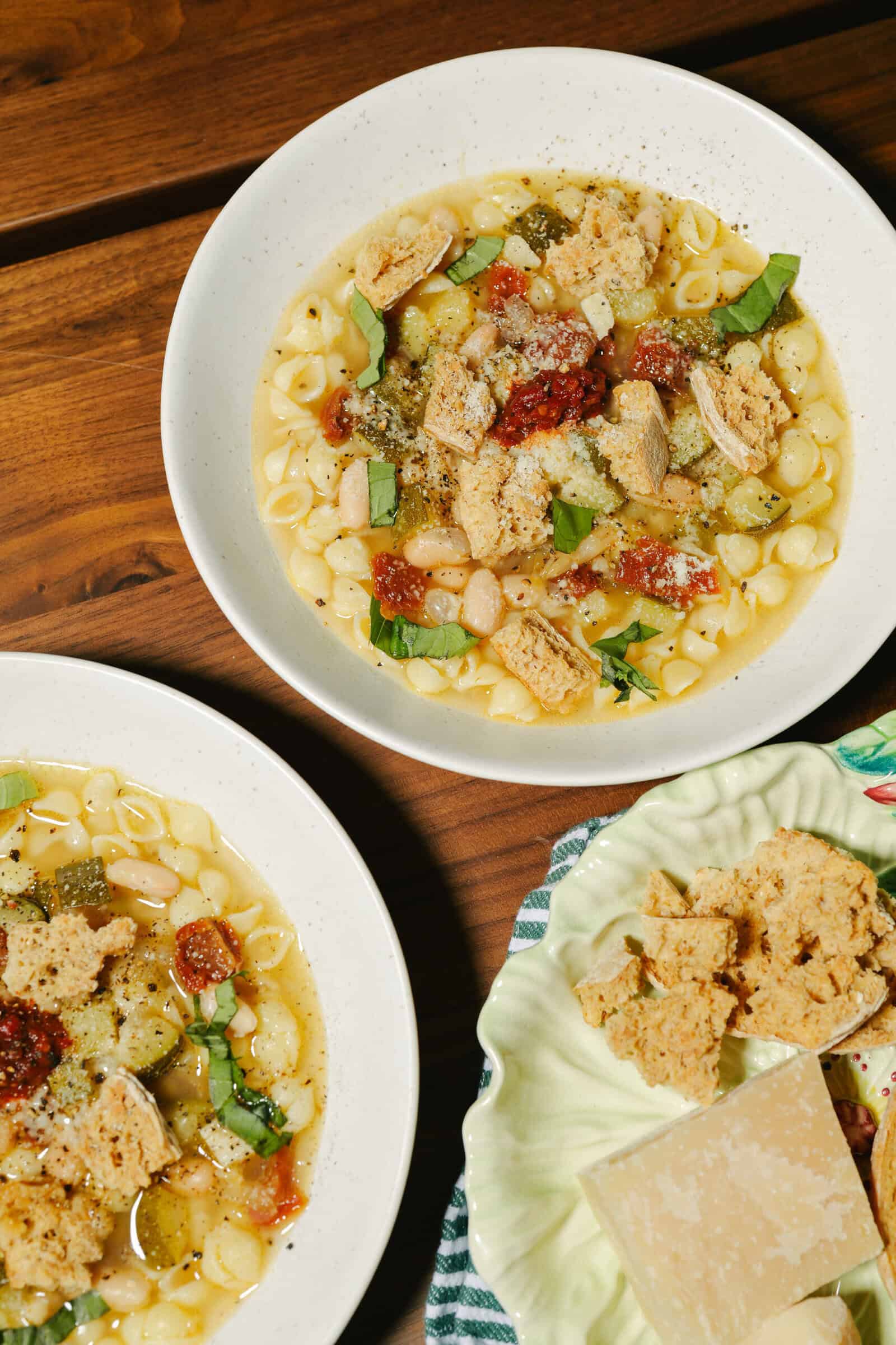Two bowls of pasta soup with white beans, tomatoes, fresh basil, crumbled crackers, and grated cheese are on a wooden table. A small plate with extra crackers and cheese is also visible.