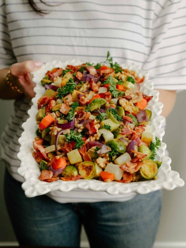 A person in a striped shirt holds a large white platter filled with a colorful roasted vegetable medley, including Brussels sprouts, potatoes, red onions, carrots, and garnished with fresh parsley.