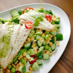 A close-up of grilled fish fillets served on a fresh salad with corn, edamame, cucumbers, and sliced red chili, as green dressing is being poured over the dish on a white oval plate.