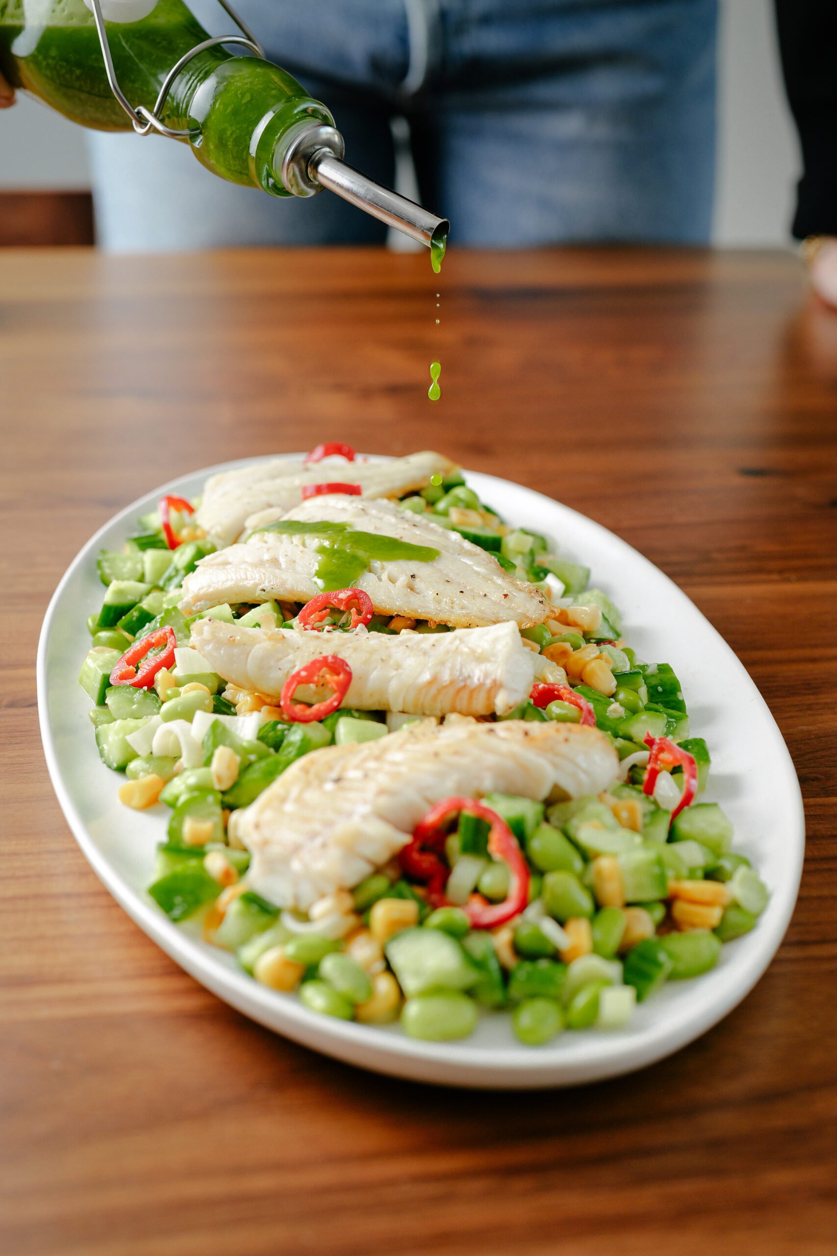 A person pours green herb oil over a salad with pieces of grilled fish, sliced red chili, corn, and diced vegetables on a white oval plate, set on a wooden table.