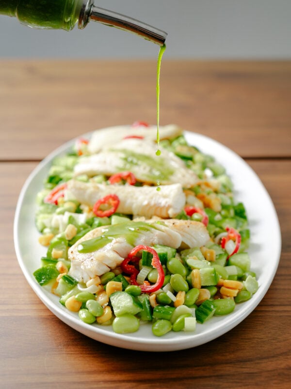 A close-up of green dressing being drizzled over a plated salad with white fish, lima beans, corn, sliced red chili peppers, and green vegetables on a white oval dish.