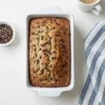 A loaf of Brown Butter Banana Bread in a white baking dish sits next to a bowl of chocolate chips, a cup of coffee, and a blue-and-white striped kitchen towel on a white surface.