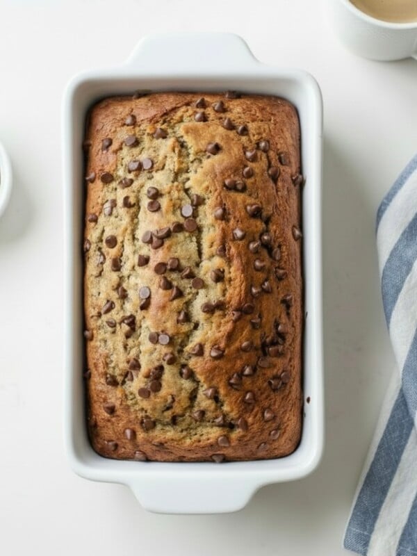 A loaf of Brown Butter Banana Bread in a white baking dish sits next to a bowl of chocolate chips, a cup of coffee, and a blue-and-white striped kitchen towel on a white surface.