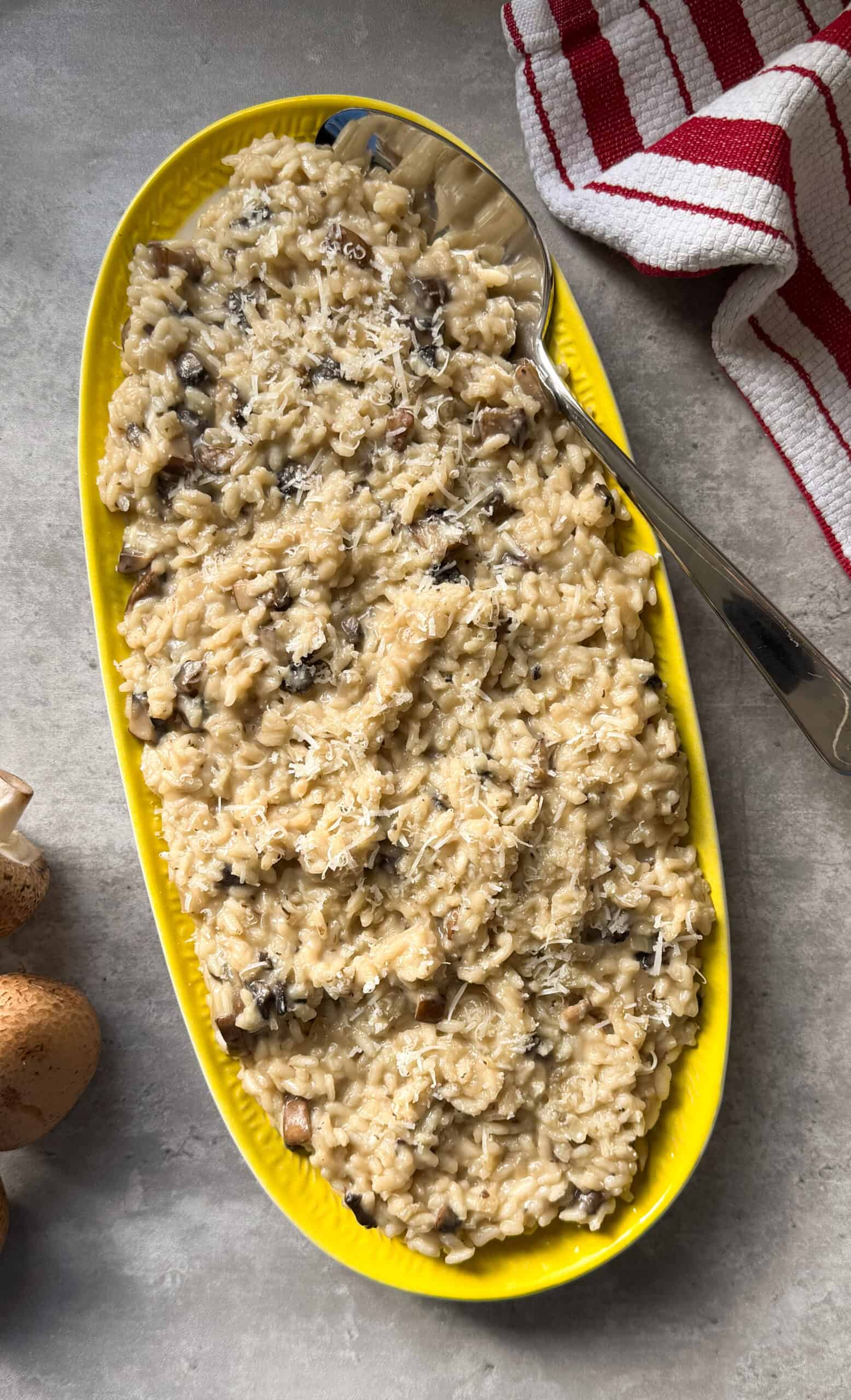 A yellow oval dish filled with creamy Mushroom Risotto, topped with grated cheese, sits on a gray surface. A large serving spoon rests in the dish, and a red-and-white striped towel is nearby&mdash;perfect for an easy risotto recipe.