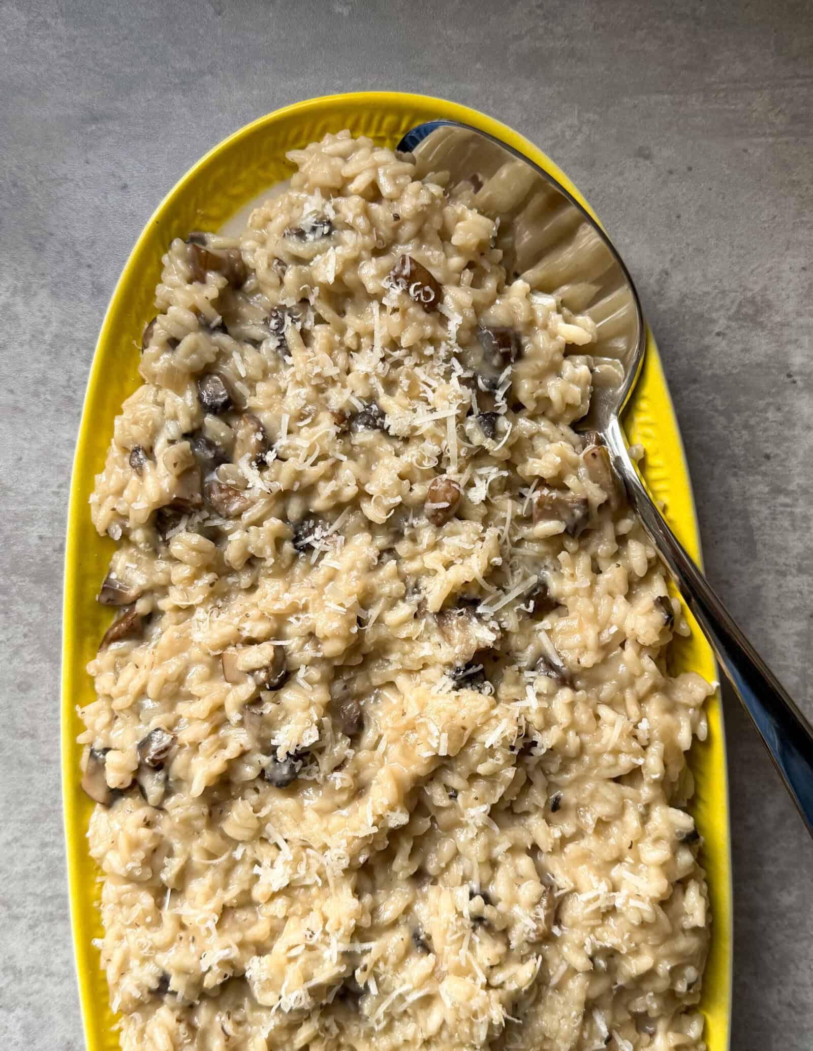 A bright yellow oval dish filled with creamy Mushroom Risotto, topped with grated cheese, sits on a gray surface with a large serving spoon resting on the side&mdash;perfect for trying this Easy Risotto Recipe at home.
