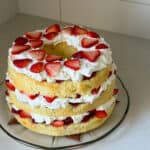 A three-layer Chiffon Cake with whipped cream and sliced strawberries between each layer and on top, displayed on a glass plate on a light-colored countertop—a delightful take on the classic Strawberry Shortcake Recipe.