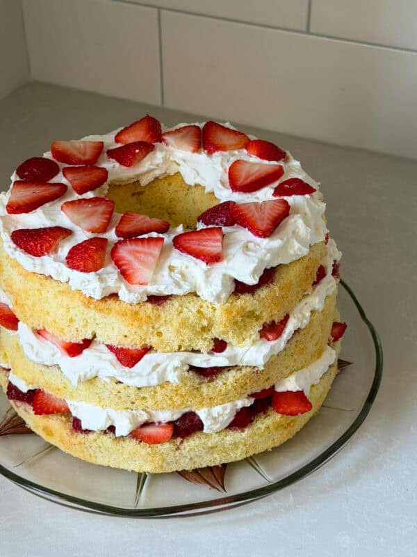 A three-layer Chiffon Cake with whipped cream and sliced strawberries between each layer and on top, displayed on a glass plate on a light-colored countertop—a delightful take on the classic Strawberry Shortcake Recipe.