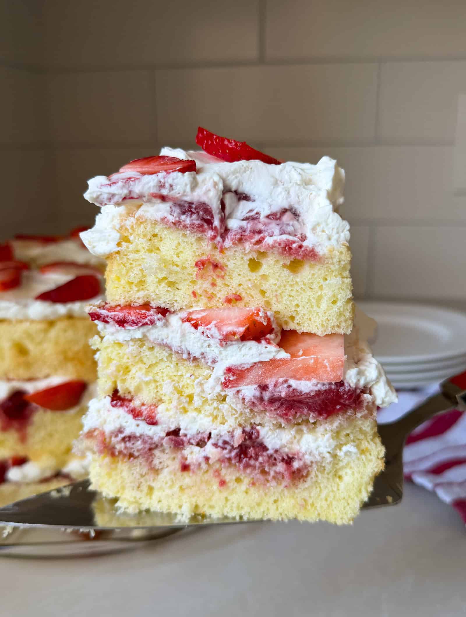 A close-up of a slice of layered chiffon cake with whipped cream and fresh strawberry slices, set against a kitchen background with stacked white plates—perfect inspiration for your next Chiffon Cake Recipe.