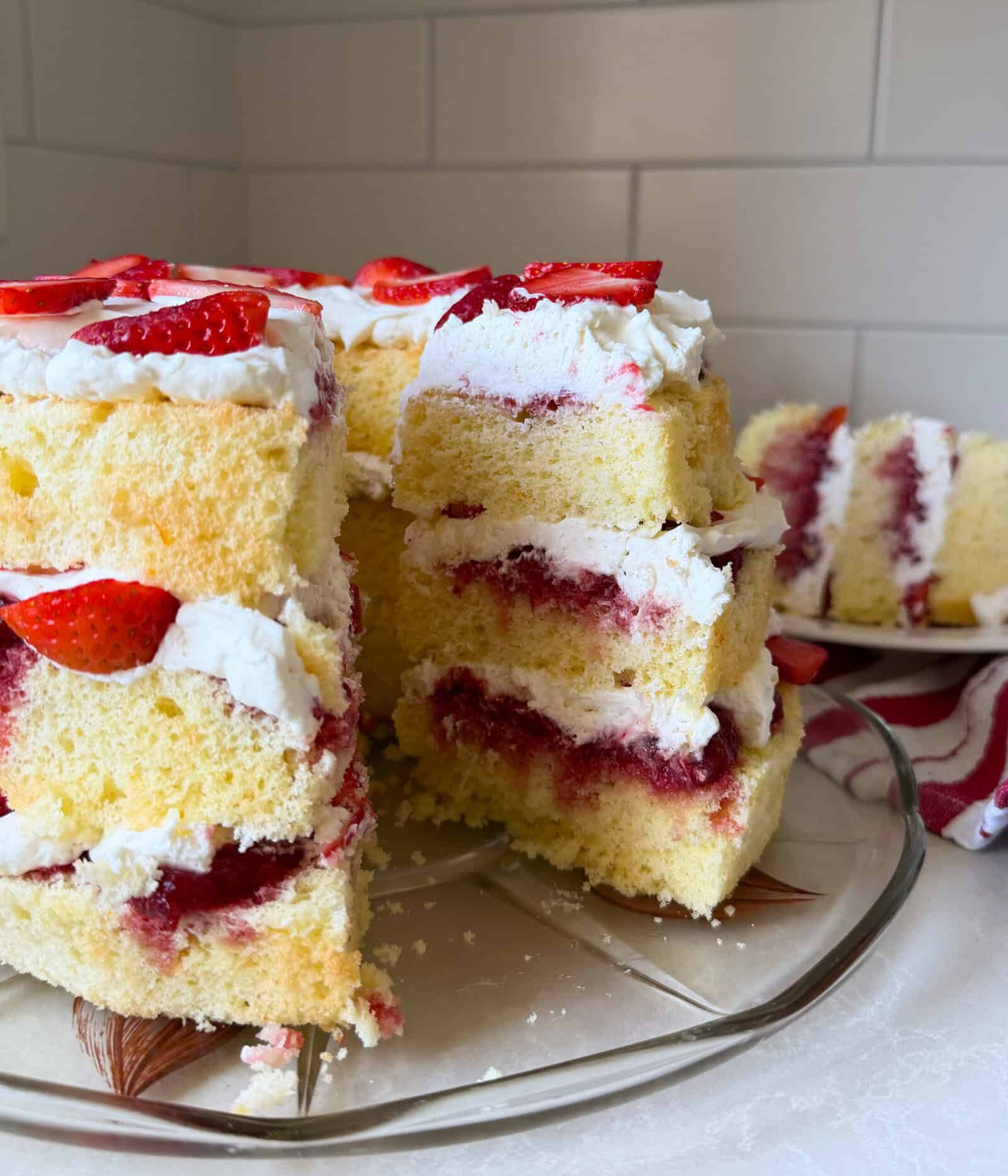 A classic Strawberry Shortcake with whipped cream and strawberry jam filling sits on a glass plate, topped with fresh strawberries and a large slice cut out. Perfect for anyone searching for the ultimate Strawberry Shortcake Recipe. White tiled wall in the background.