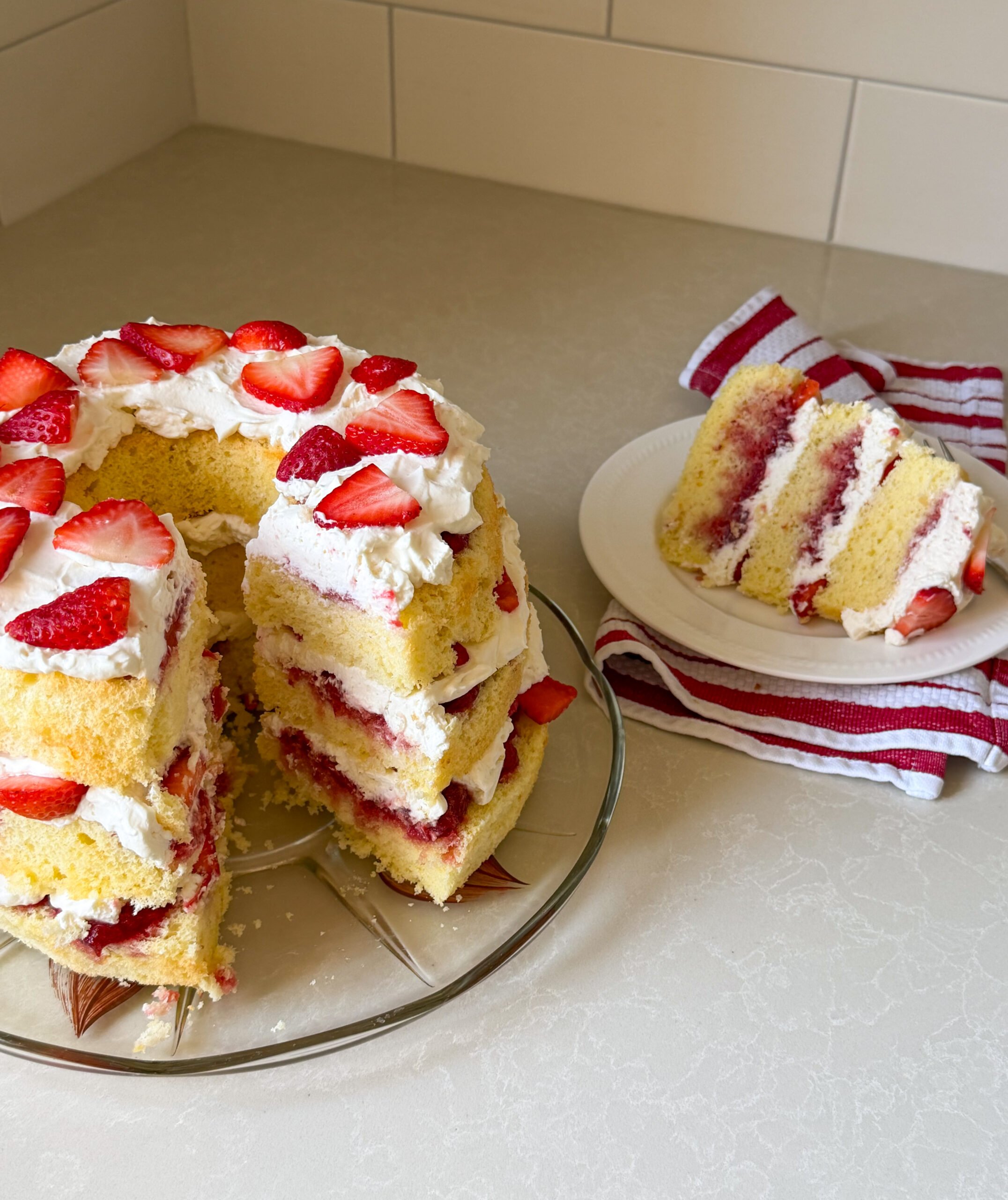 A sliced Strawberry Shortcake with layers of whipped cream and strawberries on a glass plate. A piece of this delightful Chiffon Cake is served on a white plate next to a red-striped napkin on a light kitchen countertop.