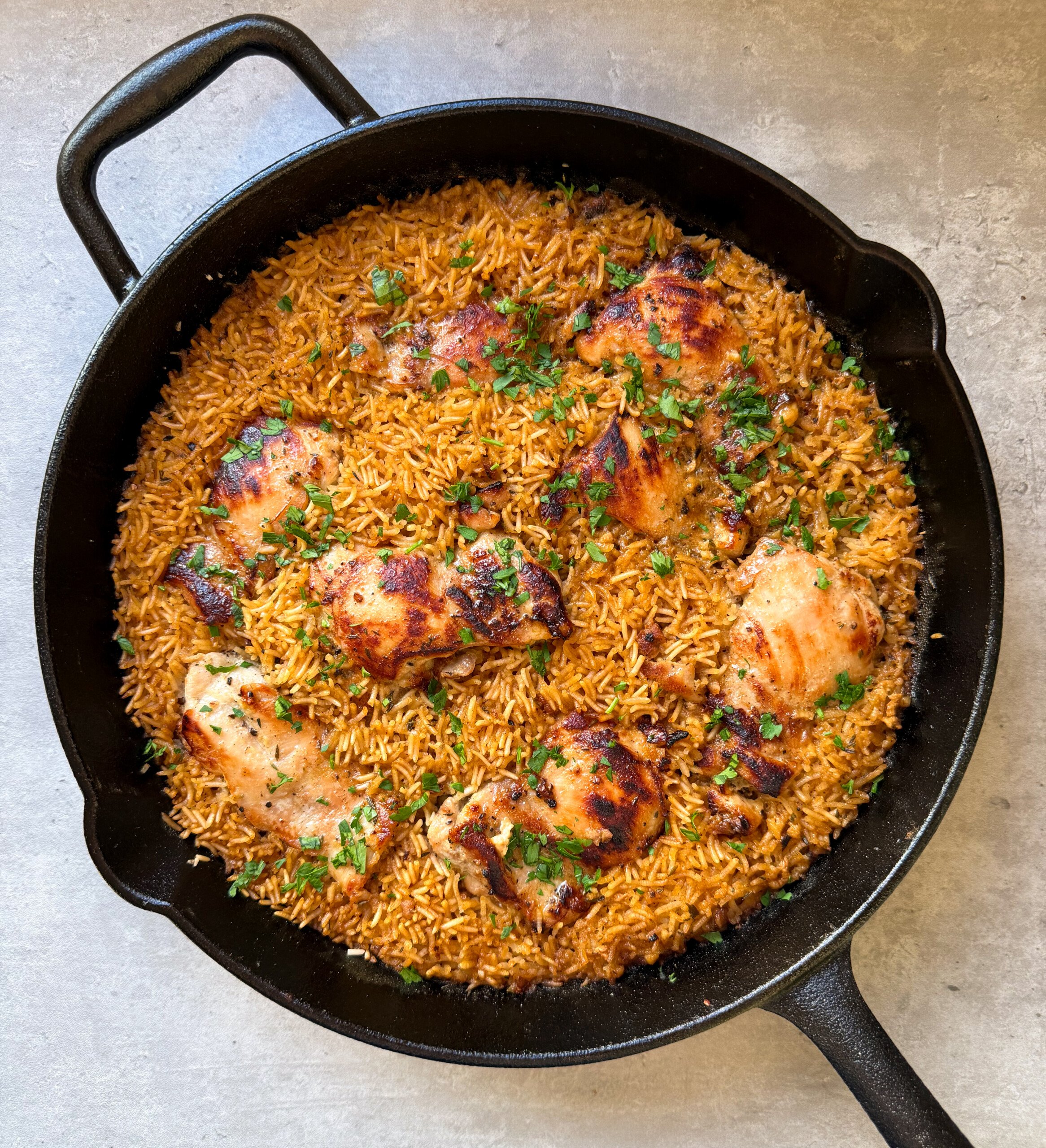 A cast iron skillet filled with golden-brown, honey mustard chicken thighs and rice, garnished with chopped herbs, sits on a light surface.