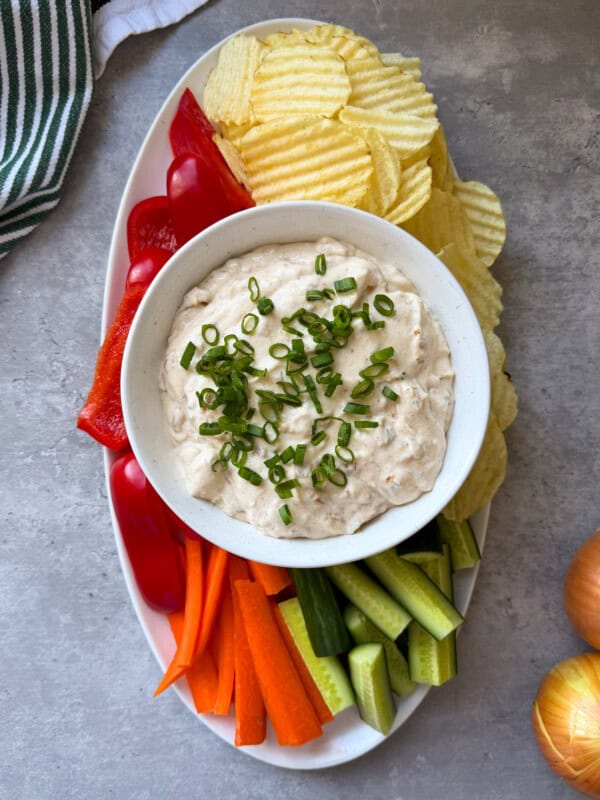 A platter with a bowl of creamy Whipped Cottage Cheese Dip topped with chopped green onions, surrounded by potato chips, red bell pepper slices, carrot sticks, and cucumber sticks. A striped towel and onions are nearby on a gray surface.