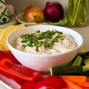 A bowl of creamy Whipped Onion Cottage Cheese Dip is garnished with chopped green onions, surrounded by sliced red bell peppers, cucumbers, carrots, and potato chips. Onions, celery, and other vegetables appear in the background.