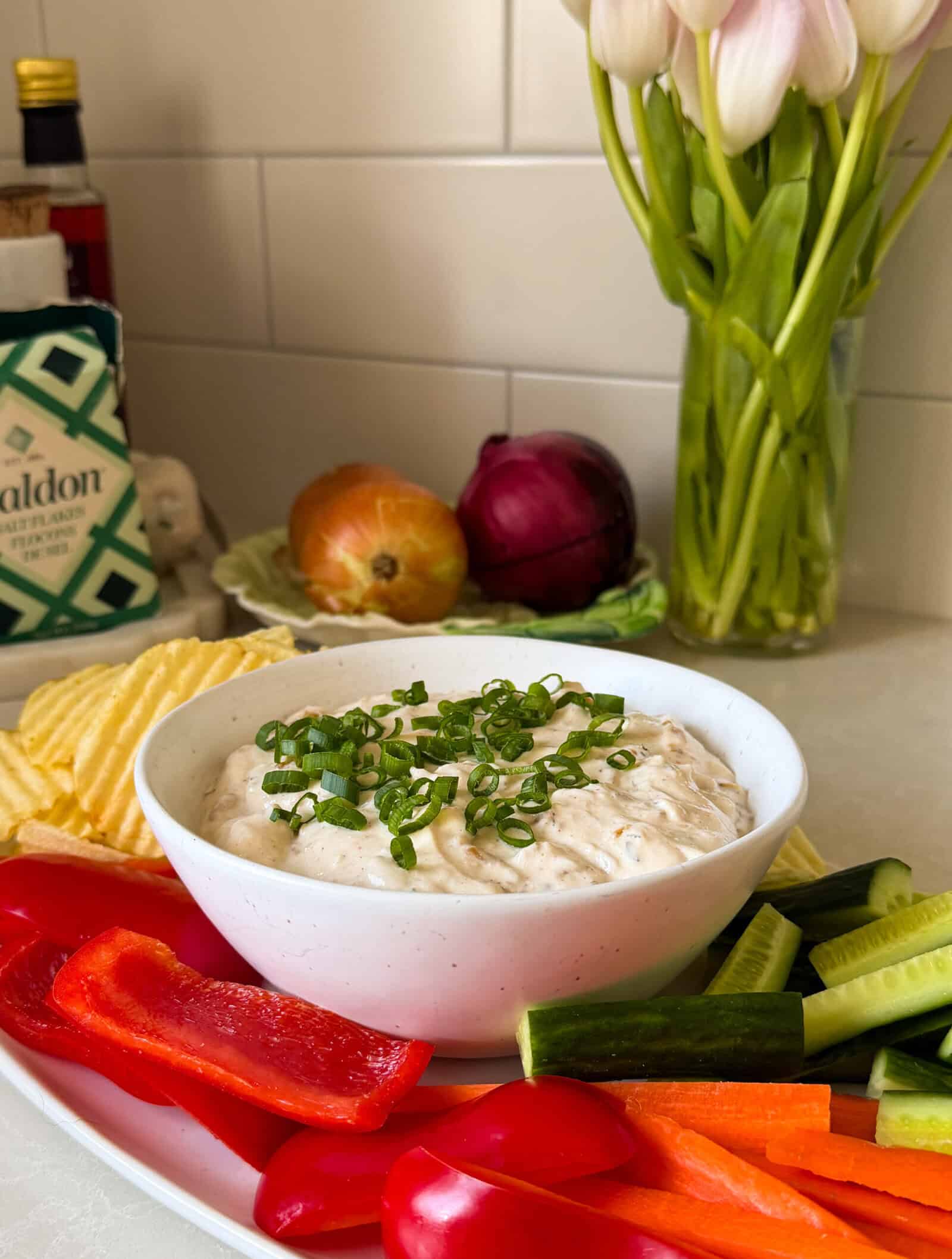 A bowl of creamy Cottage Cheese Dip topped with chopped green onions is surrounded by sliced red bell peppers, cucumbers, grape tomatoes, and potato chips. In the background are an onion, red onion, a salt box, and a vase of tulips.