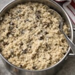 A pot of creamy Mushroom Risotto, made with this easy risotto recipe, is topped with grated cheese and a metal serving spoon. A red and white striped kitchen towel rests beside the pot on a gray countertop.