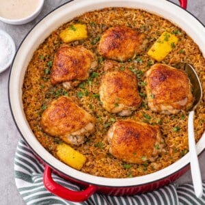 A red pot filled with golden-brown chicken thighs on a bed of seasoned rice, garnished with parsley and lemon wedges, with a serving spoon inside. A green-striped towel and small bowls are nearby.