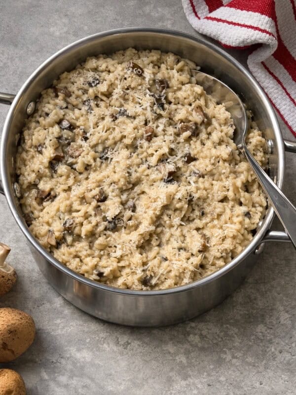 A stainless steel pot filled with creamy Mushroom Risotto sits on a gray countertop, topped with grated cheese. A metal serving spoon rests in the pot, and a red-striped towel and whole mushrooms are nearby for this Easy Risotto Recipe.