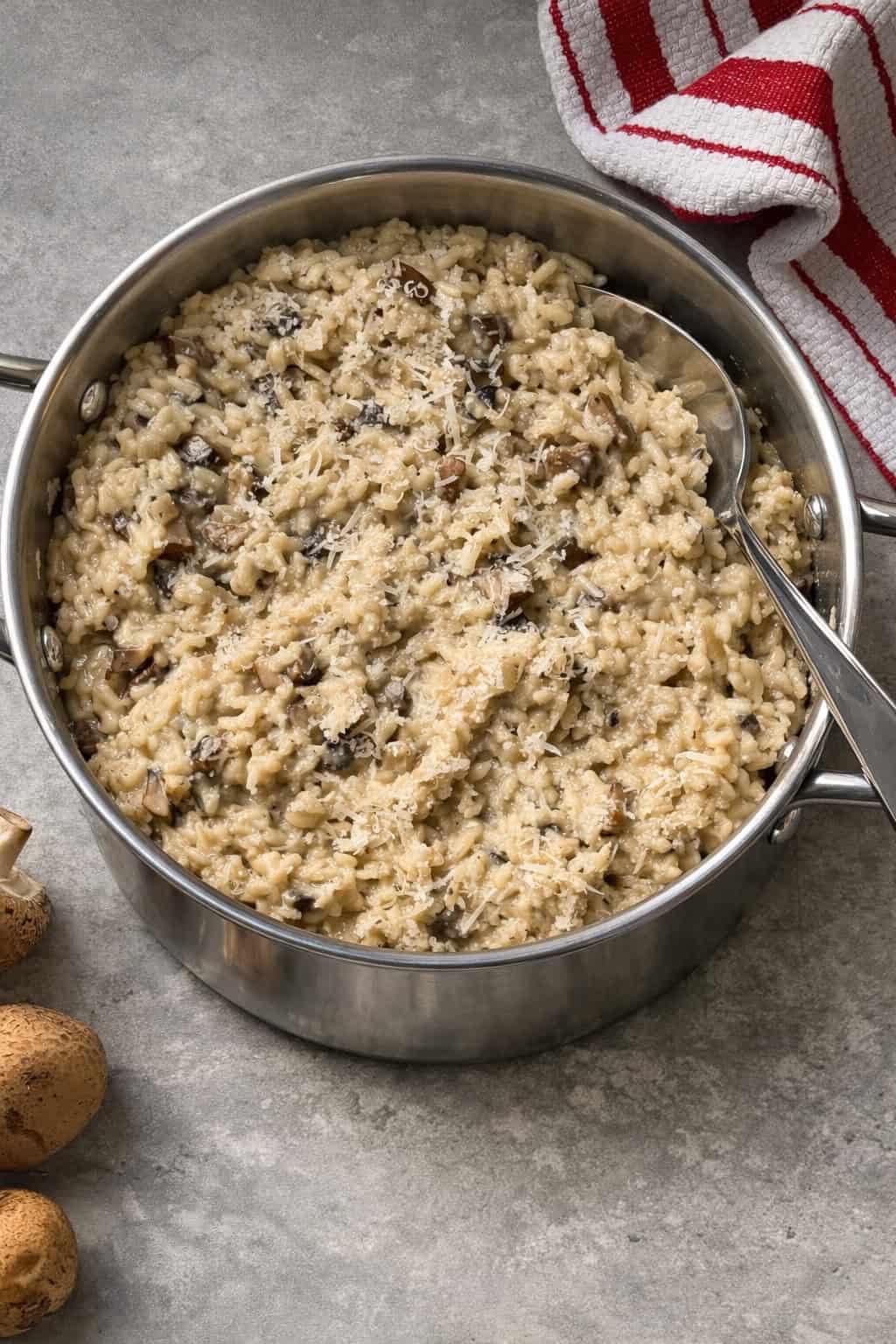A stainless steel pot filled with creamy Mushroom Risotto sits on a gray countertop, topped with grated cheese. A metal serving spoon rests in the pot, and a red-striped towel and whole mushrooms are nearby for this Easy Risotto Recipe.