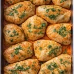 A baking dish filled with twelve stuffed cabbage rolls, inspired by Cabbage Recipes, topped with chopped parsley and sitting in a reddish sauce. The rolls are neatly arranged and baked to a golden brown.