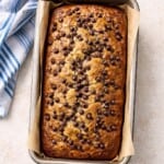 A freshly baked Banana Chocolate Chip Bread sits in a parchment-lined metal pan on a light surface, topped with chocolate chips and placed next to a blue and white striped kitchen towel.