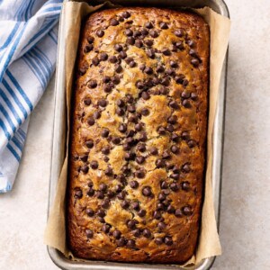 A freshly baked Banana Chocolate Chip Bread sits in a parchment-lined metal pan on a light surface, topped with chocolate chips and placed next to a blue and white striped kitchen towel.