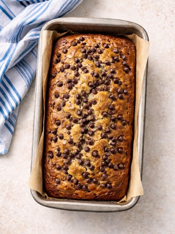 A freshly baked Banana Chocolate Chip Bread sits in a parchment-lined metal pan on a light surface, topped with chocolate chips and placed next to a blue and white striped kitchen towel.