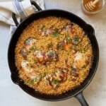 A black skillet filled with baked orzo and browned honey mustard chicken thighs, garnished with chopped herbs, sits on a light surface next to a jar of honey with a dipper and a folded kitchen towel.