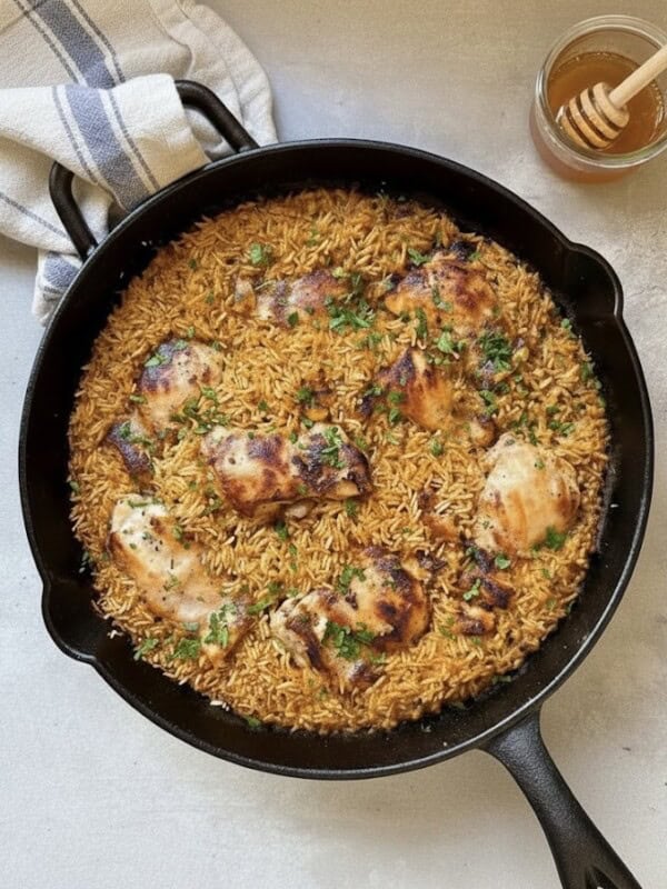 A black skillet filled with baked orzo and browned honey mustard chicken thighs, garnished with chopped herbs, sits on a light surface next to a jar of honey with a dipper and a folded kitchen towel.