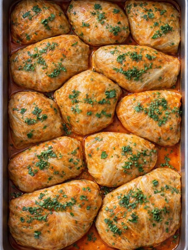 A baking dish filled with twelve stuffed cabbage rolls, inspired by Cabbage Recipes, topped with chopped parsley and sitting in a reddish sauce. The rolls are neatly arranged and baked to a golden brown.