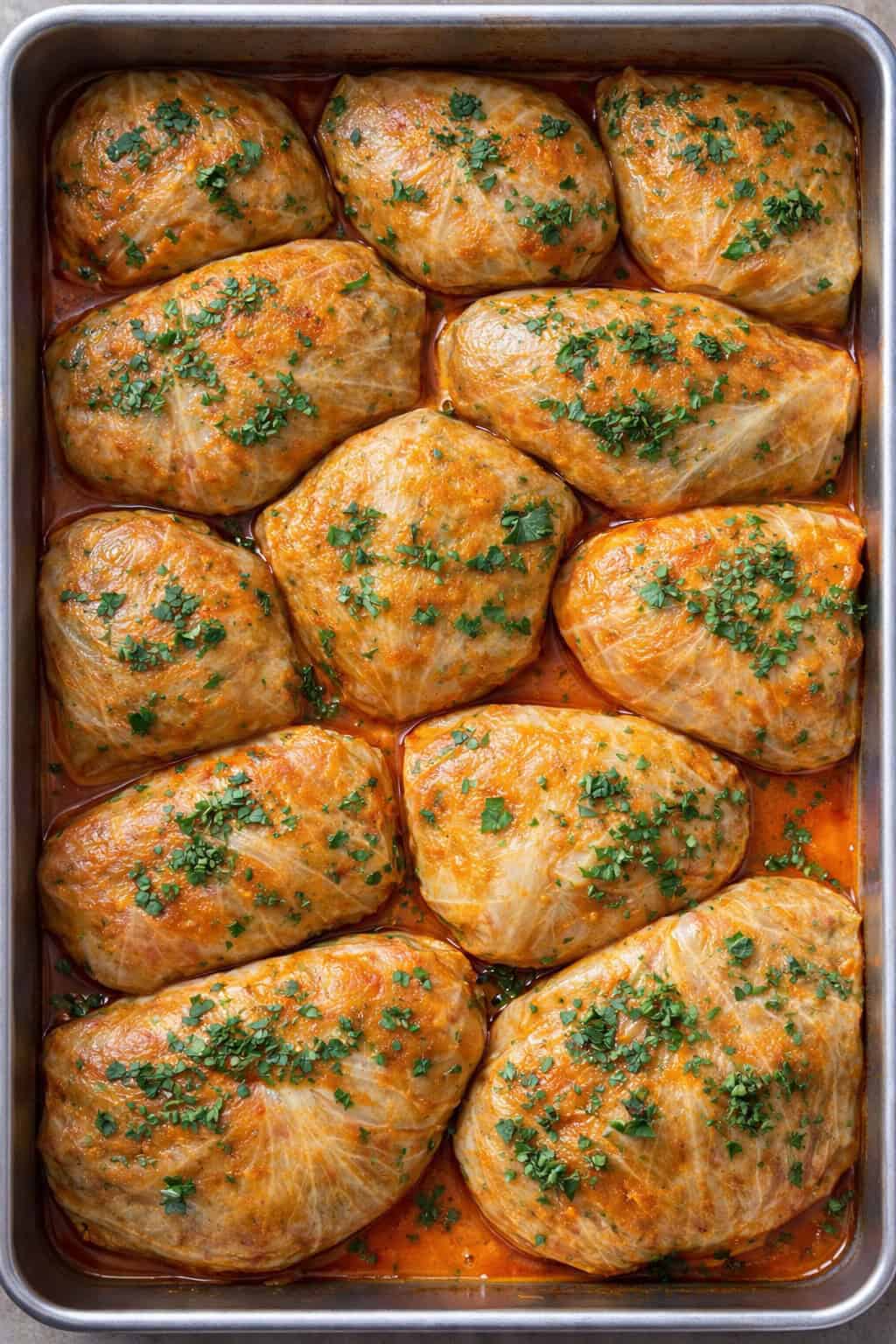 A baking dish filled with twelve stuffed cabbage rolls, inspired by Cabbage Recipes, topped with chopped parsley and sitting in a reddish sauce. The rolls are neatly arranged and baked to a golden brown.