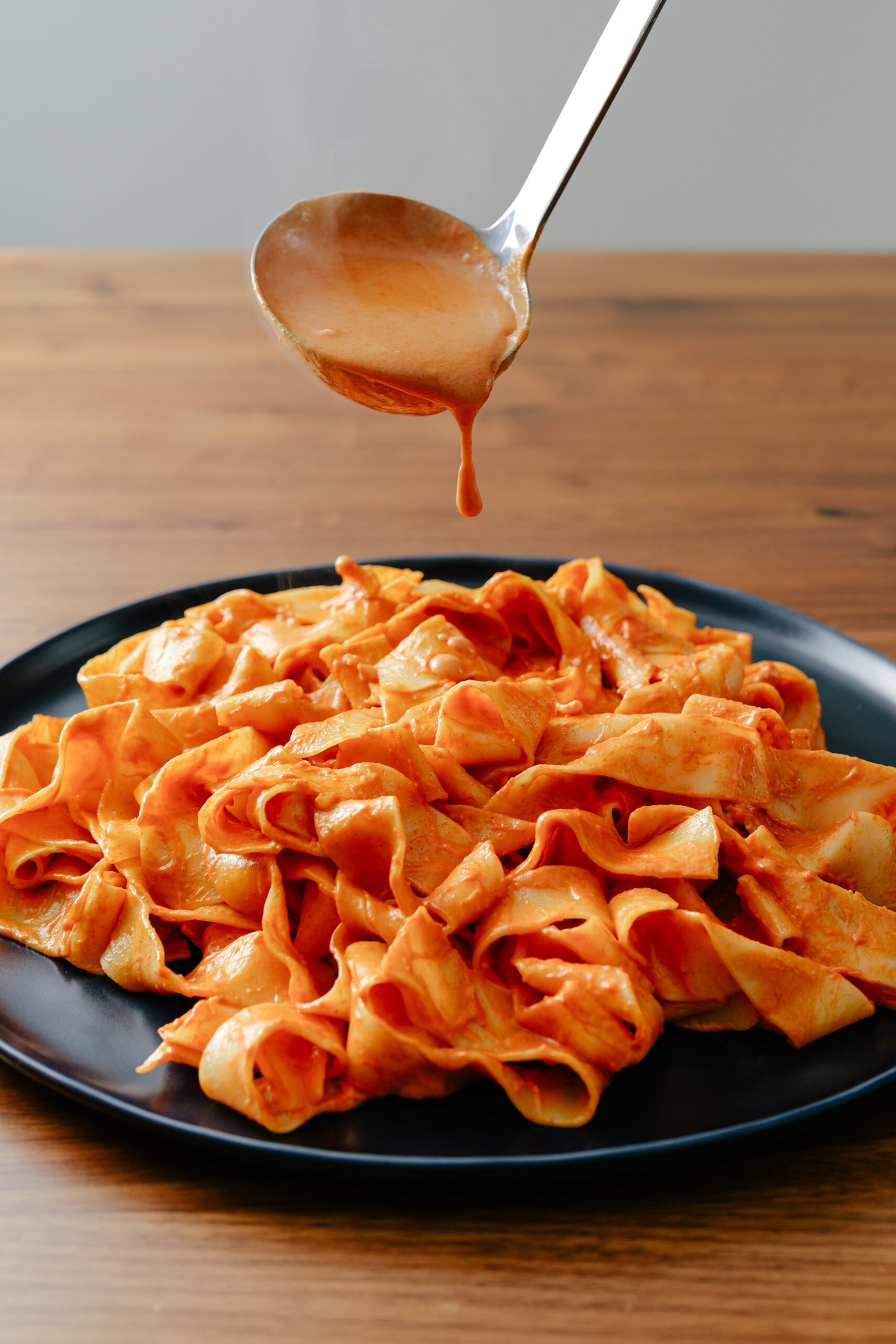 A black plate filled with wide, flat pasta noodles coated in red tomato sauce sits on a wooden table. A ladle hovers above, dripping extra sauce onto the pasta.