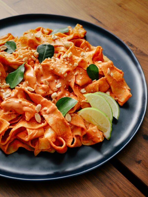 A black plate of wide, flat pasta covered in a creamy orange sauce, garnished with fresh green leaves, chopped peanuts, and lime wedges, on a wooden table.