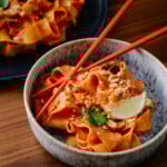 A bowl of wide, flat noodles in a red sauce, garnished with crushed peanuts, lime wedge, and green herbs. Red chopsticks rest on the bowl. Another plate of noodles is in the background on a wooden table.