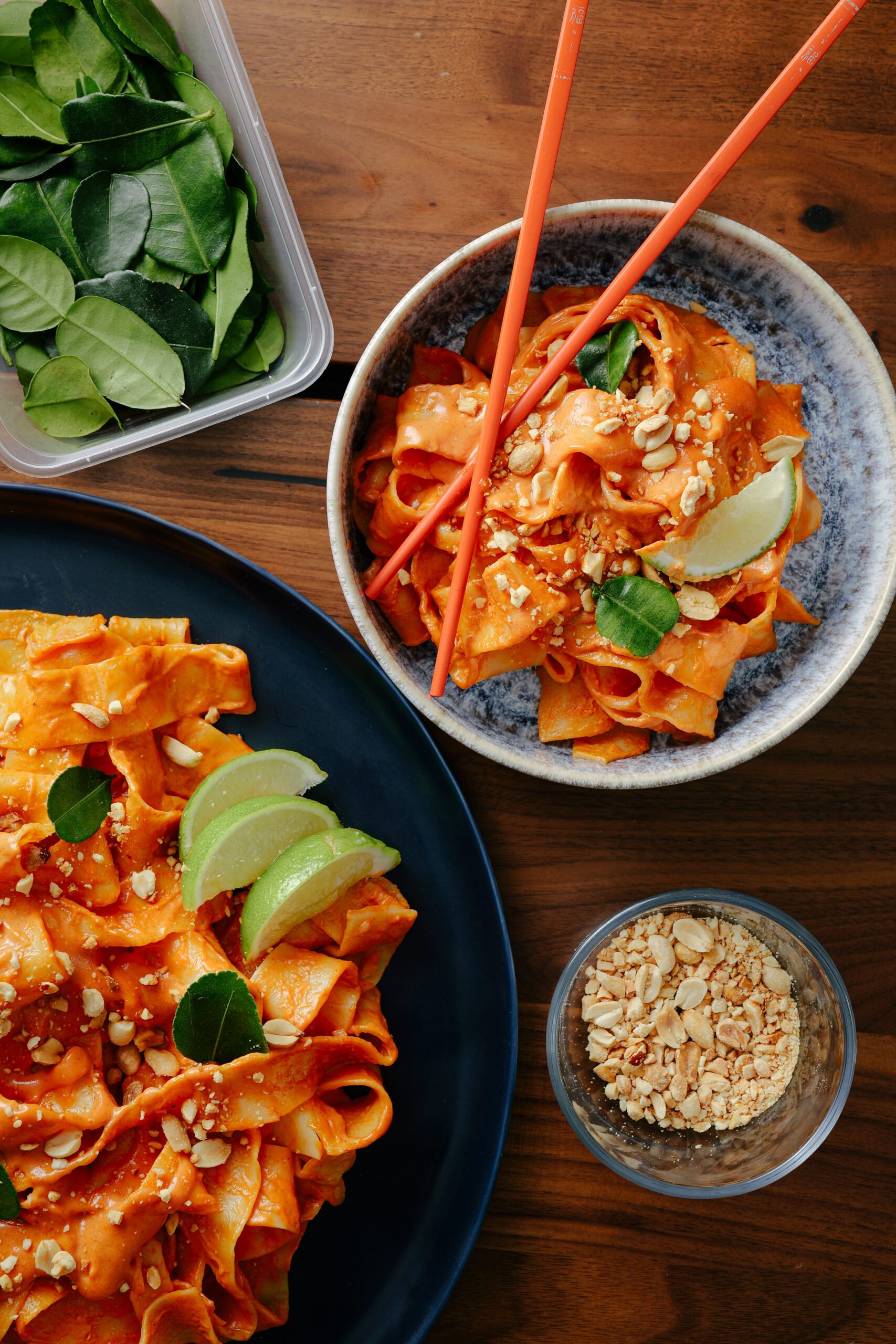 A bowl of wide, flat noodles in red sauce, garnished with lime wedges, peanuts, and green leaves, sits beside a plate of more noodles, a container of green leaves, and a small bowl of chopped peanuts on a wooden table.