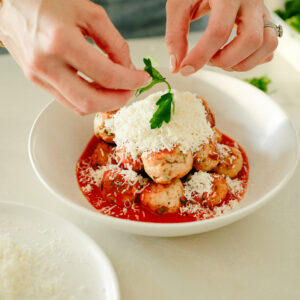 A person garnishes a bowl of meatballs in tomato sauce with fresh parsley and grated cheese, using their hands over the dish on a light-colored countertop.