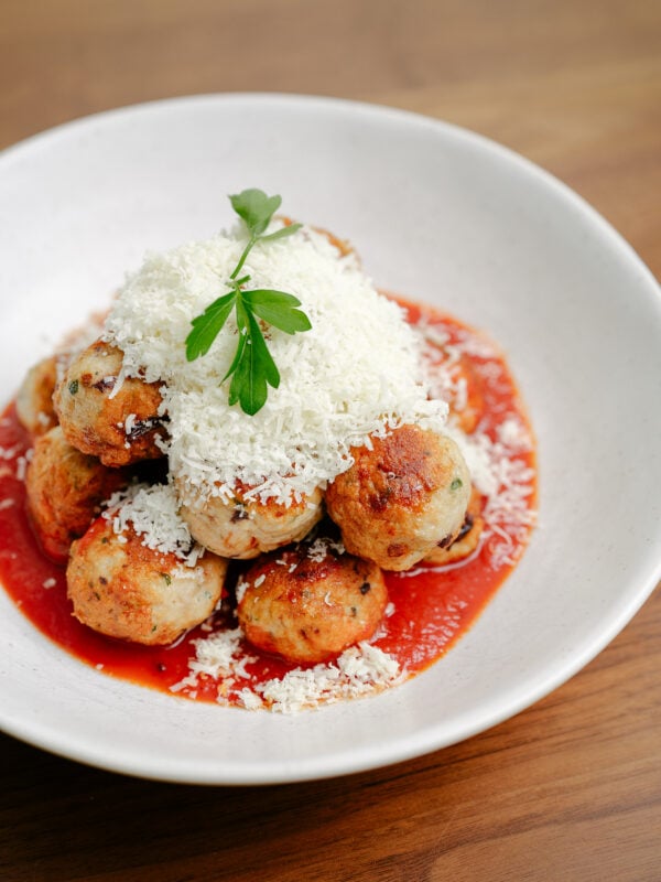 A white bowl filled with meatballs in red tomato sauce, topped with a generous amount of grated cheese and garnished with a sprig of fresh parsley, sits on a wooden table.