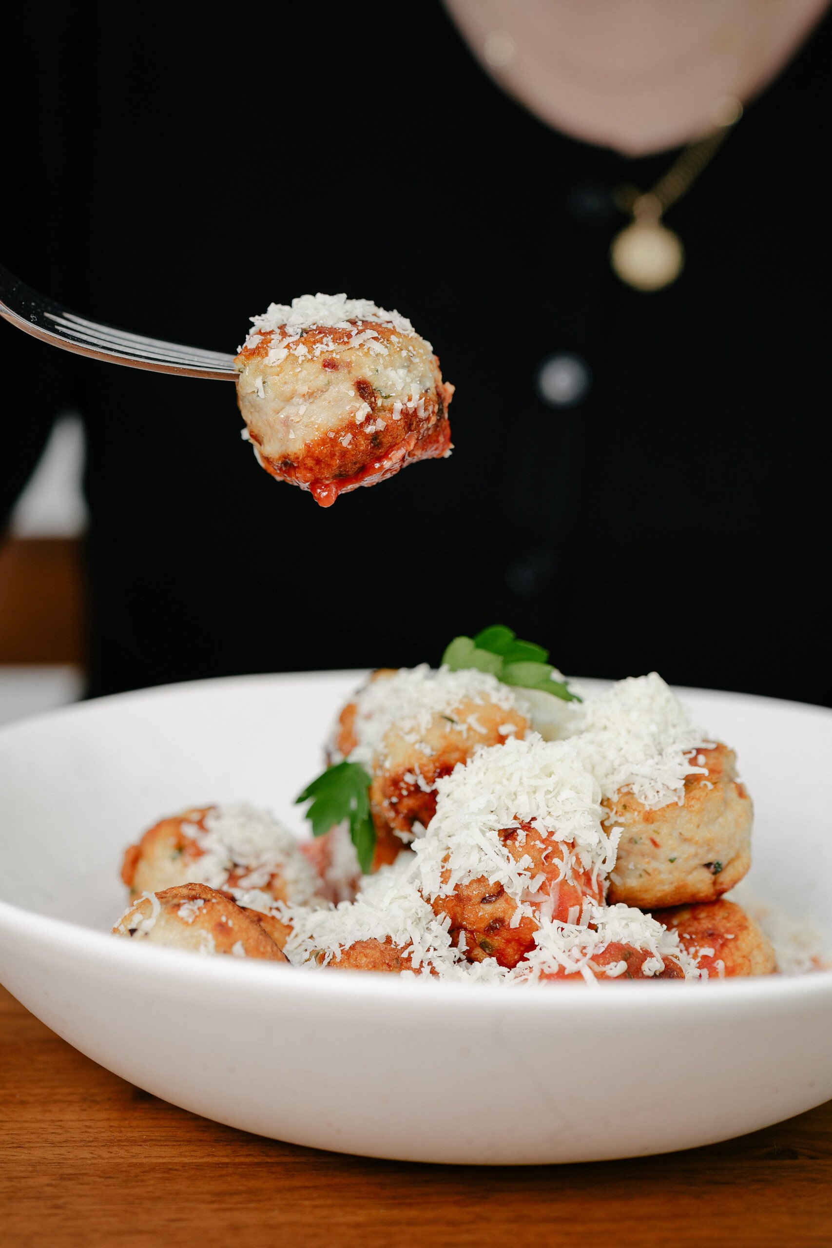 A person holds a fork with a meatball topped with grated cheese above a bowl filled with meatballs, tomato sauce, parsley, and more grated cheese.