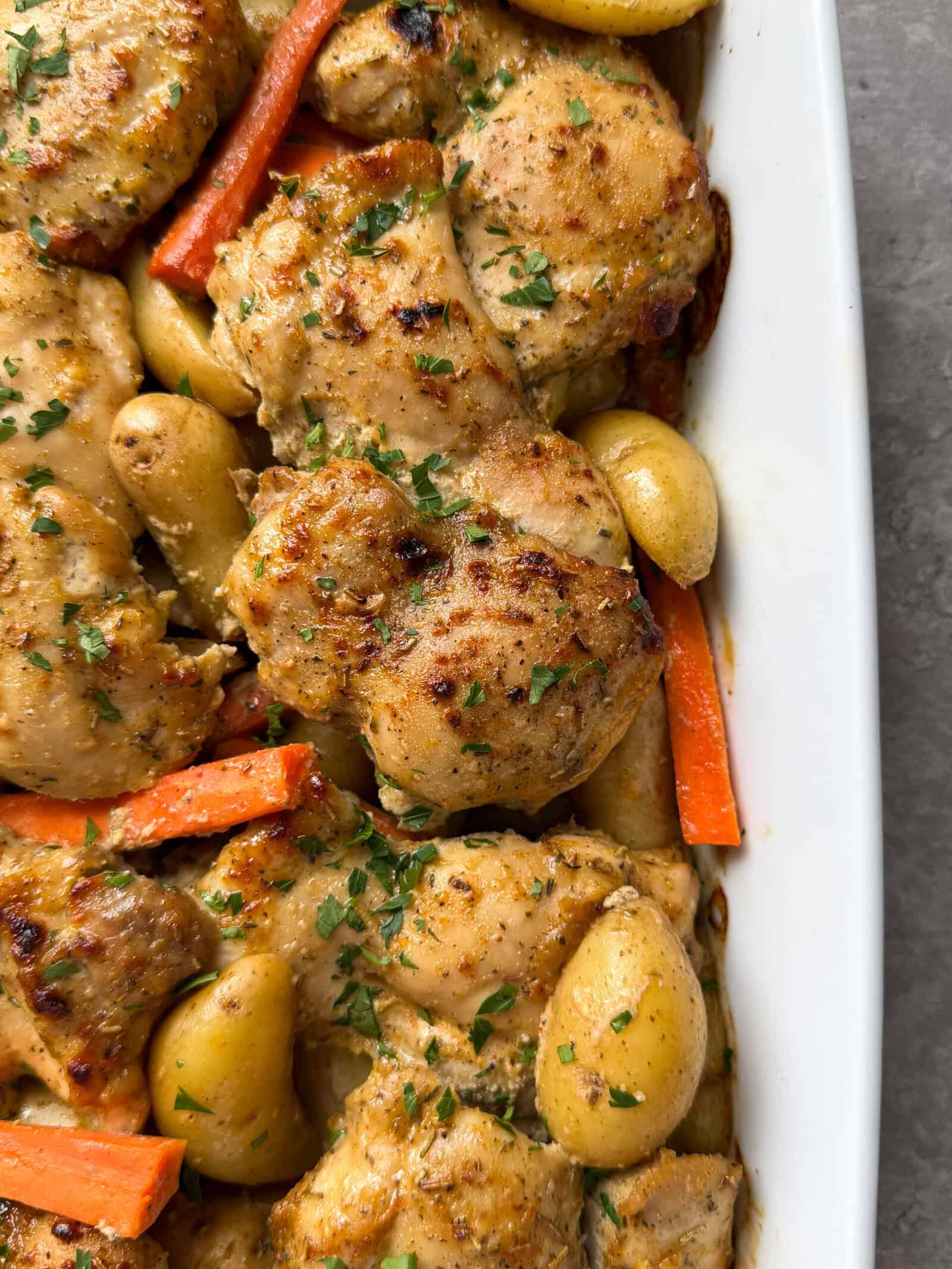 Baked chicken thighs with herbs, baby potatoes, and carrot sticks arranged in a white baking dish make a tasty chicken sheet pan dinner. The dish is garnished with chopped parsley for a perfect finishing touch.