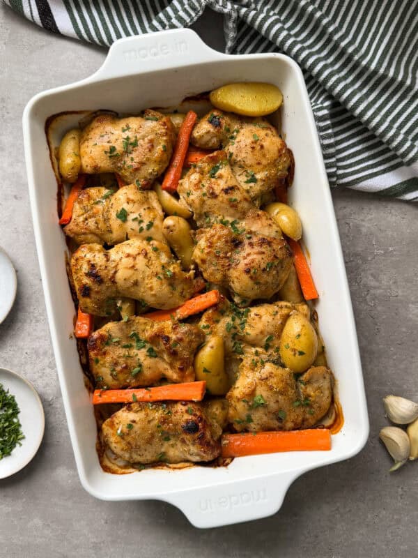 A white baking dish filled with roasted chicken thighs, carrots, and small potatoesโan inviting chicken sheet pan dinnerโgarnished with herbs. A green and white striped towel and a few garlic cloves rest nearby on a grey surface.