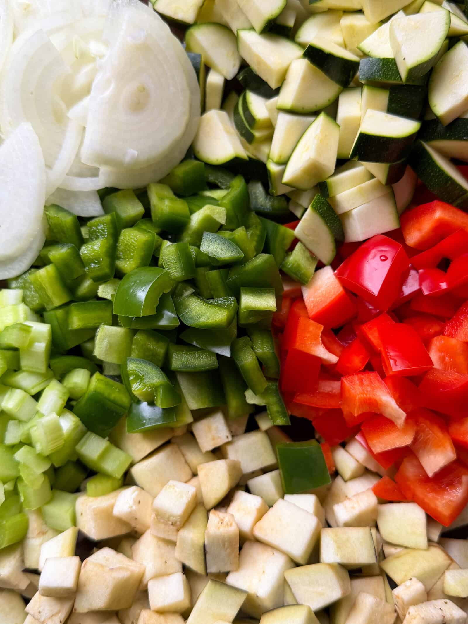 A close-up of chopped vegetables, including white onion slices, green bell pepper, red bell pepper, celery, zucchini, and eggplant—classic ingredients for a vibrant Caponata recipe—arranged in a colorful, organized pattern.