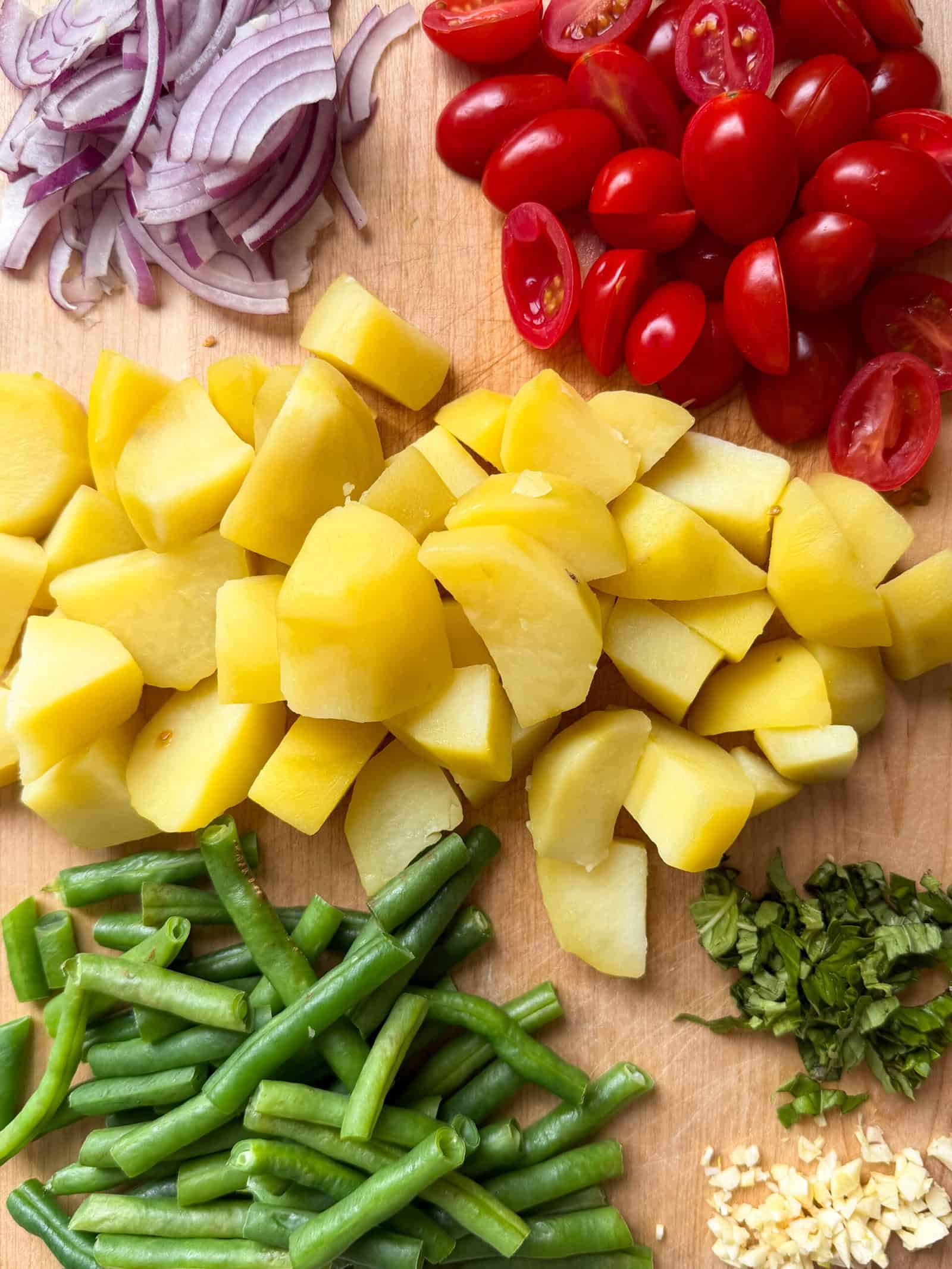 Chopped red onions, halved cherry tomatoes, diced potatoes, green beans, minced garlic, and chopped herbs are arranged in separate sections on a wooden board for an Italian Potato Salad Recipe.