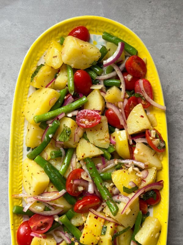 A vibrant Italian Potato Salad with green beans, cherry tomatoes, red onion, and herbs, served on a yellow platter against a gray background. The vegetables are mixed and lightly dressed with seasoning.