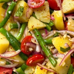 A close-up of a vibrant Italian Potato Salad with potatoes, green beans, cherry tomatoes, red onion slices, and herbs, sprinkled with red pepper flakes and vinaigrette.