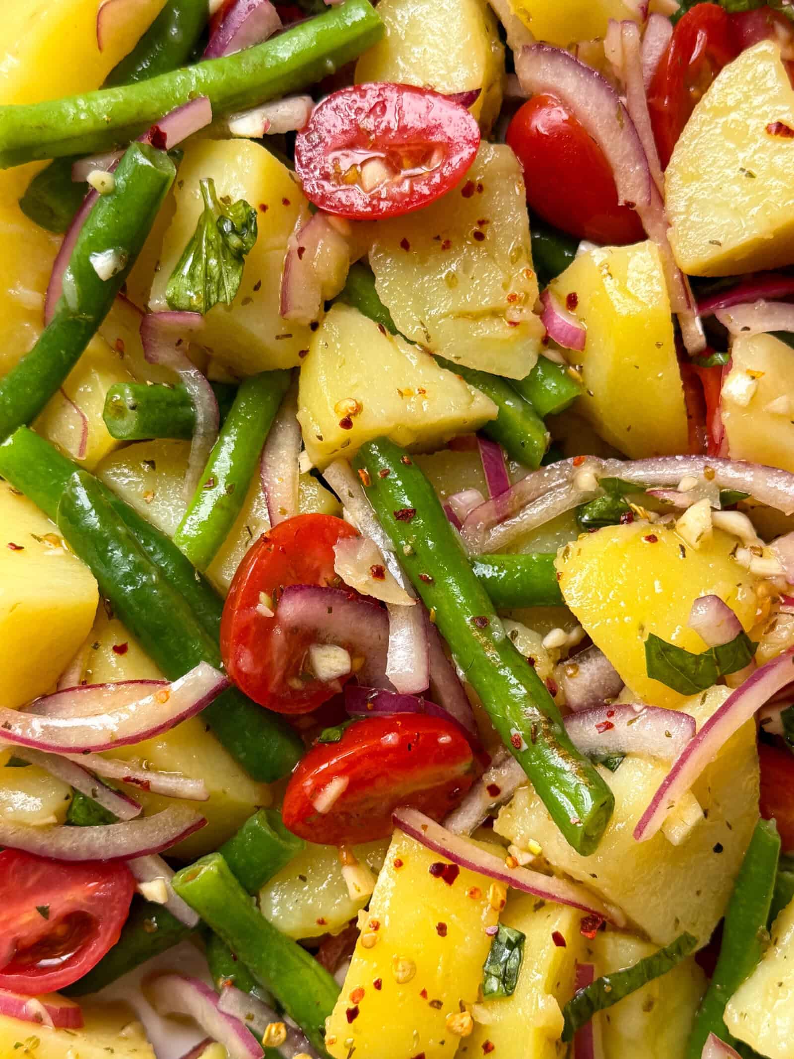 A close-up of a vibrant Italian Potato Salad with potatoes, green beans, cherry tomatoes, red onion slices, and herbs, sprinkled with red pepper flakes and vinaigrette.