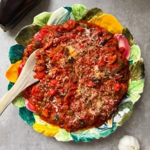 A colorful leaf-shaped plate filled with ratatouille sits on a gray surface, topped with grated cheese. A wooden spoon rests in the dish, evoking a Caponata vibe. Nearby are a whole eggplant and two bulbs of garlic.