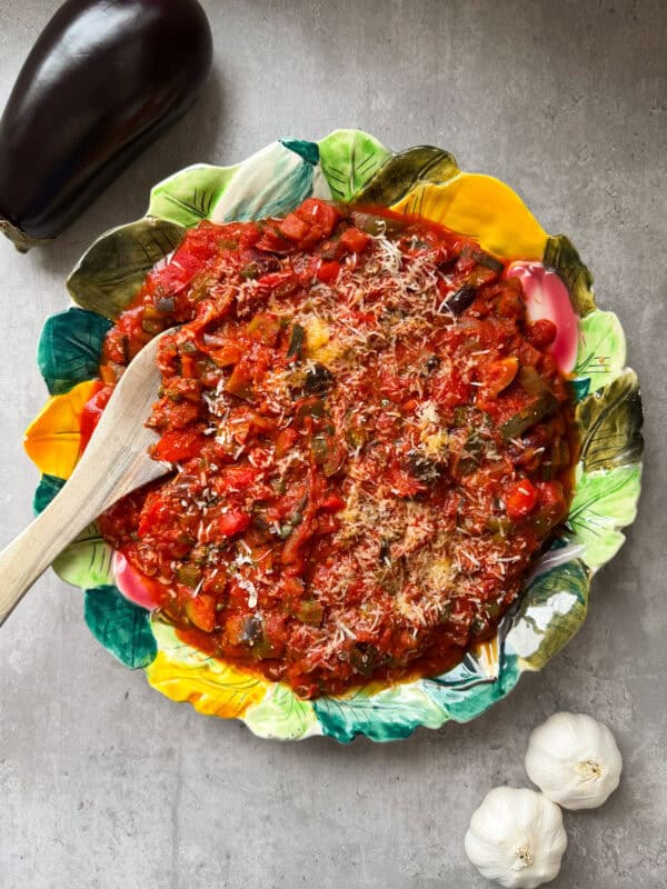 A colorful leaf-shaped plate filled with ratatouille sits on a gray surface, topped with grated cheese. A wooden spoon rests in the dish, evoking a Caponata vibe. Nearby are a whole eggplant and two bulbs of garlic.